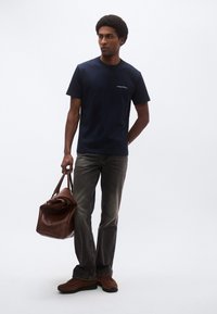 Tengerészkék pamutpóló kerek nyakkal, szürkésfakó farmernadrággal, barna szarvasbőr cipővel és barna bőr utazótáskával párosítva.