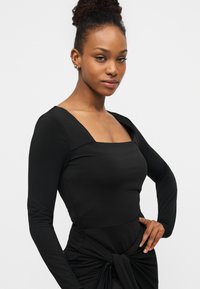 Μαύρο μακρυμάνικο τοπ με τετράγωνη λαιμόκοψη και εφαρμοστή σχεδίαση. Διαθέτει λεπτομέρεια με κόμπο στη μέση και έχει απαλή, ελαστική υφή.