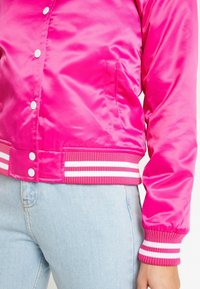 Pink satin bomber jacket with white snap buttons and striped ribbed cuffs and hem. Features a side pocket and smooth texture.