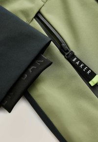 Grøn og sort jakke med en lynlåslomme med sort hardware og et mærke mærket "BAKER." Glat stof med et tekstureret underlag.