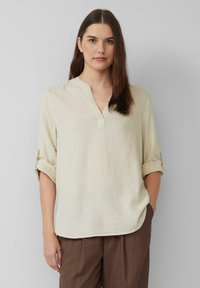 Beige, leichte Leinenbluse mit V-Ausschnitt, hochgekrempelten Ärmeln und lockerer Passform, kombiniert mit braunen Hosen. Einfaches Design.