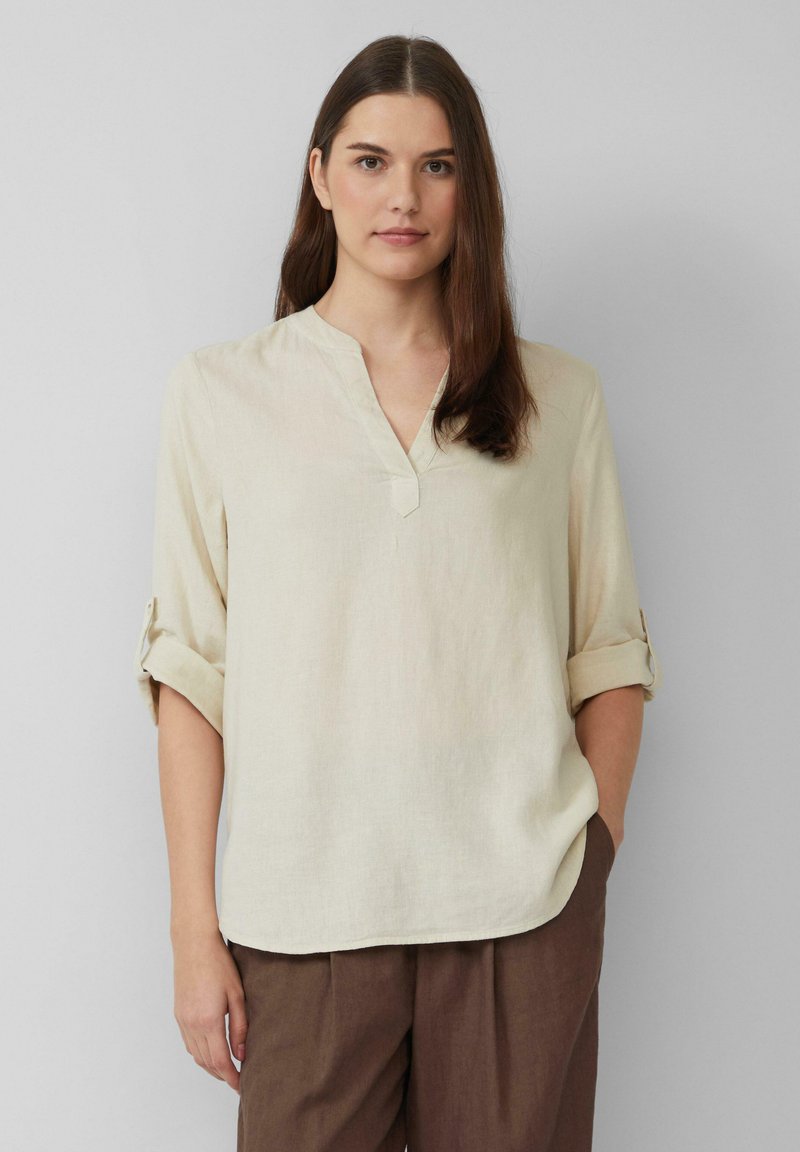 Beige, leichte Leinenbluse mit V-Ausschnitt, hochgekrempelten Ärmeln und lockerer Passform, kombiniert mit braunen Hosen. Einfaches Design.