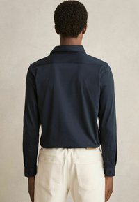 Cămașă lungă navy, cu o textură moale, având guler ascuțit și manșete cu nasturi, purtată cu pantaloni off-white, prezentând un fit relaxat.