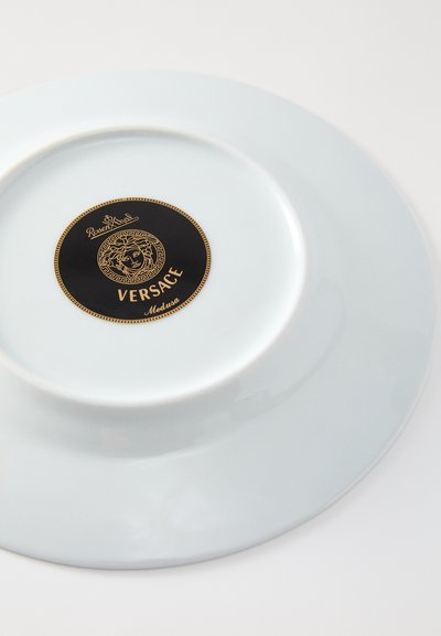 Biała ceramiczna talerz z błyszczącym wykończeniem, z czarno-złotym logo z motywem Meduzy na środku. Okrągły kształt, gładka faktura.