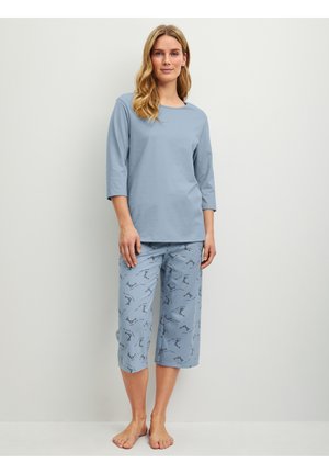 Vrouw staat blootsvoets, draagt een lichtblauw shirt met driekwart mouwen en lichtblauwe cropped broek met klein donker patroon tegen een effen muur.