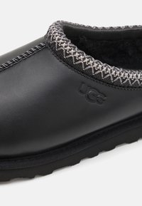 Fekete slip-on cipő sima bőr felsőrésszel, szürke hímzett varrással a gallér mentén és texturált gumitalppal. Az UGG márkajelzés jellemzi.