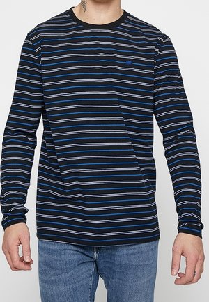 Mężczyzna ubrany w czarną koszulę z długim rękawem z poziomymi niebiesko-białymi paskami, w niebieskich dżinsach, stojący na jednolitym tle.