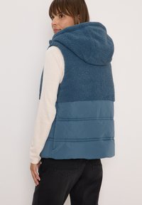 Blaues Weste mit einem pelzartigen Oberteil und glattem Unterteil, mit Kapuze und gestepptem Muster. Wird über einem hellen Langarmshirt getragen.