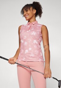Sleeveless pink polo with white floral pattern, collared design, and logo detail at neckline. Held golf club, wearing matching pink pants.