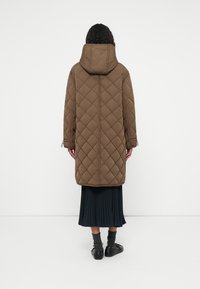 Casaco castanho acolchoado com um design de diamante, capuz preso, cordões laterais e um corte até ao joelho, combinado com uma saia preta plissada.