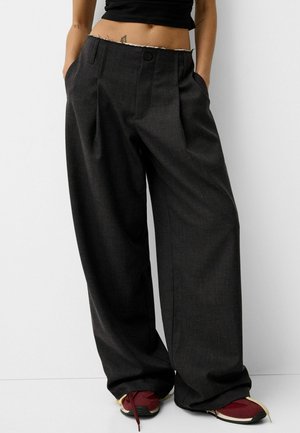 Osoba w za dużych, ciemnoszarych plisowanych spodniach, czarnym krótkim topie i czerwonych trampkach ze sznurowadłami rozwiązanymi, stojąca z rękami w kieszeniach.