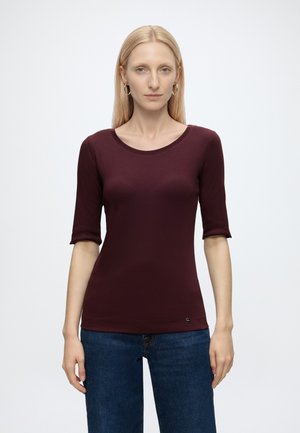 Femme aux longs cheveux blonds portant un haut bordeaux ajusté à manches courtes et un jean bleu, debout devant un fond clair uni.