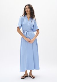 Hellblaues, knielanges Wickelkleid mit kurzen Ärmeln und passendem Bindegürtel. Glattes Stoff mit einer subtilen Textur. Schwarze Flip-Flops getragen.