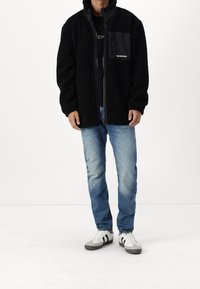 Chaqueta de forro polar negra con cremallera frontal, corte oversize, bolsillos laterales y un bolsillo en el pecho con un logo. Combinada con vaqueros azules y zapatillas.