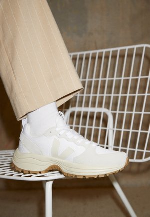 Fod med hvid sneakers med beige sål, hvid strømpe og beige bukser med nålestriber, hvilende på hvid trådstol mod neutral baggrund.