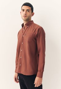 Chemise marron boutonnée avec manches longues, une poche poitrine et une texture lisse. Elle présente un col pointu et des boutons argentés.