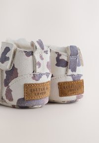 Bottes douces et texturées pour bébé en blanc avec des taches violettes et grises. Comprend une étiquette en daim beige avec l'inscription "Little & Loved" en relief. Fermetures à scratch.