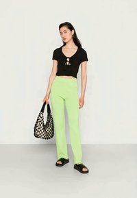 Top preto cropped com detalhe de nó à frente, combinado com calças verdes-neon de cintura alta. Modelo segura uma bolsa de rede preta e usa sandálias pretas.