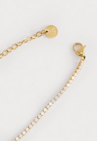 Bracciale in catena d'oro con accenti di pietre trasparenti, dotato di chiusura a gancio e un'etichetta circolare con logo. Texture liscia e finitura lucida.