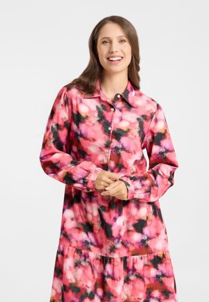 Smiling woman with long brown hair wearing a pink, black, and red abstract patterned long-sleeve button-up dress against a plain background.