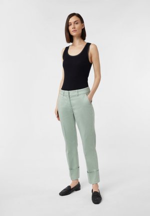 Schwarzes ärmelloses Oberteil, hellgrüne maßgeschneiderte Hose mit hochgekrempelten Säumen und schwarze Slipper. Fokussierung auf klare Linien und glatte Texturen.