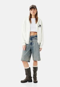 Młoda kobieta ubrana w biały krótki top, biały rozpinany hoodie, długie dżinsowe szorty, ciemne skórzane botki oraz czapkę z czarnej skóry, stoi na białym tle.