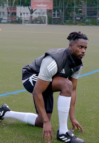 Hombre atlético negro con una chaqueta sin mangas negra y una camiseta gris, arrodillado sobre la hierba, usando calcetines blancos y zapatos de fútbol negros.