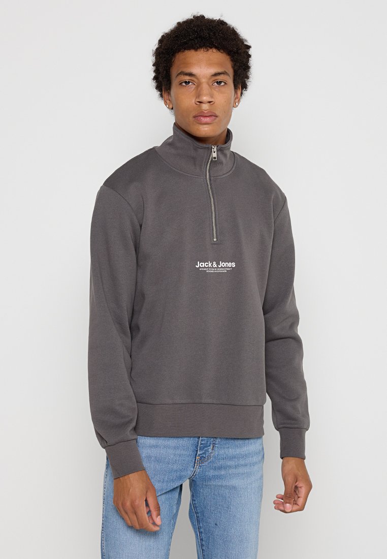 Sudadera de cremallera gris oscuro con cuello alto y dobladillo acanalado, que presenta un pequeño logotipo blanco en el pecho. Combinada con vaqueros azules.