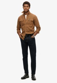 Bruine langemouwige shirt met borstzakken, gedragen over een wit T-shirt; gecombineerd met donkere jeans en zwarte chelsea boots, casual outfit.