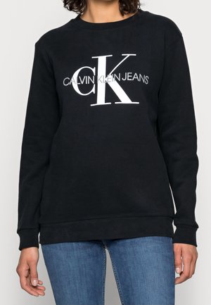 Czarna bluza z miękkiego materiału, z wyraźnym białym logo "CALVIN KLEIN JEANS" oraz okrągłym dekoltem i ściągaczami na rękawach i u dołu.
