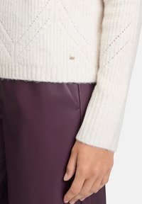 Hand ruht neben weinroten Lederhosen, trägt einen weißen gestrickten Pullover mit dezentem Muster und gerippten Bündchen.