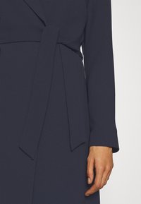 Primo piano sul lato destro di una persona che indossa un cappotto belted di un blu scuro con maniche lunghe, che mostra mano e avambraccio contro uno sfondo chiaro.