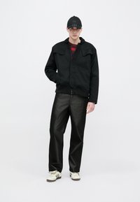 Fekete cipzáras dzseki két első zsebbel, fényes fekete bőszárú nadrággal és törtfehér sportcipővel párosítva. Fekete sapka logó részlettel.