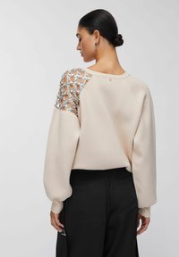 Hanorac bej deschis, cu umeri texturati și împodobiți, având un strat transparent deasupra și o croială lejeră. Mâneci cu manșete și gât rotund.