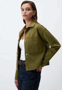 Veste cropped vert olive en tissu doux avec un col, poche avant et fermetures à pression, associée à un haut blanc et un jean noir.