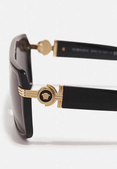 Occhiali da sole neri con una robusta montatura quadrata, dettagli in tono oro e un'emblema Medusa sulle tempie. Finitura liscia e design moderno.