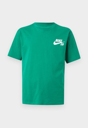 Mand stående med hænderne i lommerne iført grøn Nike SB T-shirt, sorte bukser, sorte Nike-sneakers og lyseblå kasket mod hvid baggrund.
