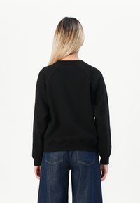 Vrouw met lang blond haar, gekleed in een zwart sweatshirt met lange mouwen en blauwe denim wijde jeans, gezien van achteren tegen een witte achtergrond.