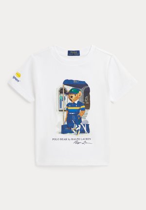 T-shirt blanc avec un ours Polo portant une casquette verte, une chemise rayée bleue et jaune, un pantalon bleu, des baskets et tenant un sac.