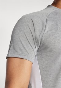 Grå treningsskjorte med rund hals, korte ermer og hvite sideakenter. Stoffet ser glatt og lett ut.