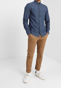 Marinblå skjorta med knappar och ett diskret mönster, kombinerad med tanfärgade smala byxor och vita sneakers. Skjortan har en bröstficka och krage.