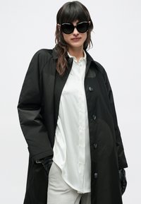 Femeie cu păr închis la culoare purtând ochelari de soare negri supradimensionați, palton negru, cămașă albă cu nasturi, pantaloni deschiși la culoare și mănuși negre, pe un fundal alb simplu.