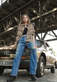 Camisa xadrez sobre uma blusa preta, jeans desfiados azuis e botins pretos. Sentado num carro castanho com uma matrícula visível. Estrutura metálica ao fundo.