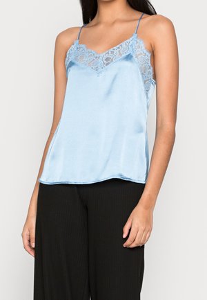 Femme portant un caraco en satin bleu clair avec des bordures en dentelle et des bretelles fines, associé à un pantalon noir ample.