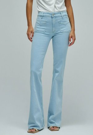 Vaqueros acampanados de tiro alto en azul claro con bolsillos frontales de parche, combinados con top blanco y sandalias beige de punta descubierta.