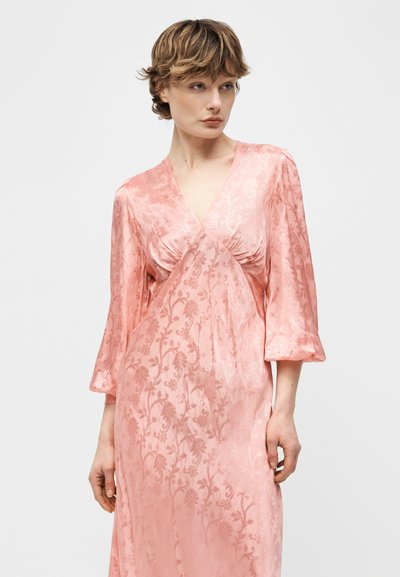 Γυναίκα που φοράει ένα ροζ, σατινέ φόρεμα με μοτίβα, μανίκια 3/4 και V-λαιμόκοψη, στέκεται μπροστά σε απλό ανοιχτό γκρι φόντο.