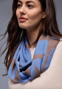 Mujer de cabello largo y castaño que lleva una bufanda con estampado azul y beige sobre una blusa beige clara, frente a un fondo gris liso.