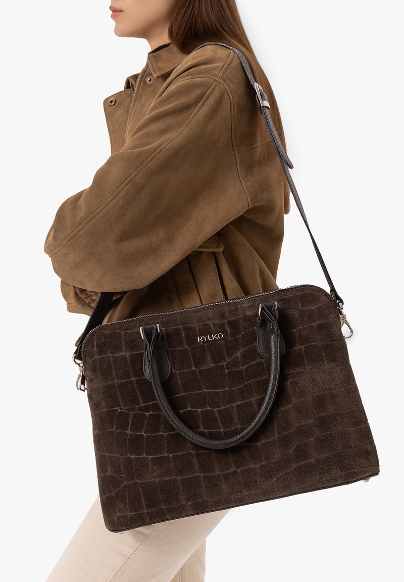 Femme portant une veste en daim marron et un sac à main marron foncé texturé avec poignées et bandoulière portée en travers du corps.
