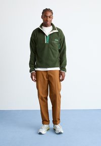 Green fleece pullover with white and green accents and a zip pocket, paired with tan trousers and gray sneakers. Smooth texture, relaxed fit.