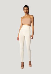 Donna con capelli ricci, che indossa un top corto strapless beige, leggings crema a vita alta e tacchi nude, in piedi davanti a uno sfondo grigio chiaro.